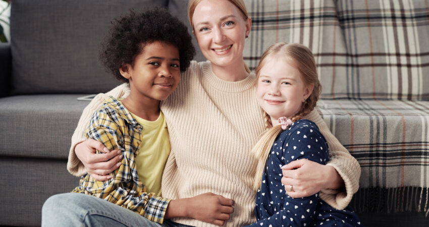 Woman sitting on the floor smiling and hugging two children, one with curly dark hair and the other with blonde hair, in a cozy living room setting.