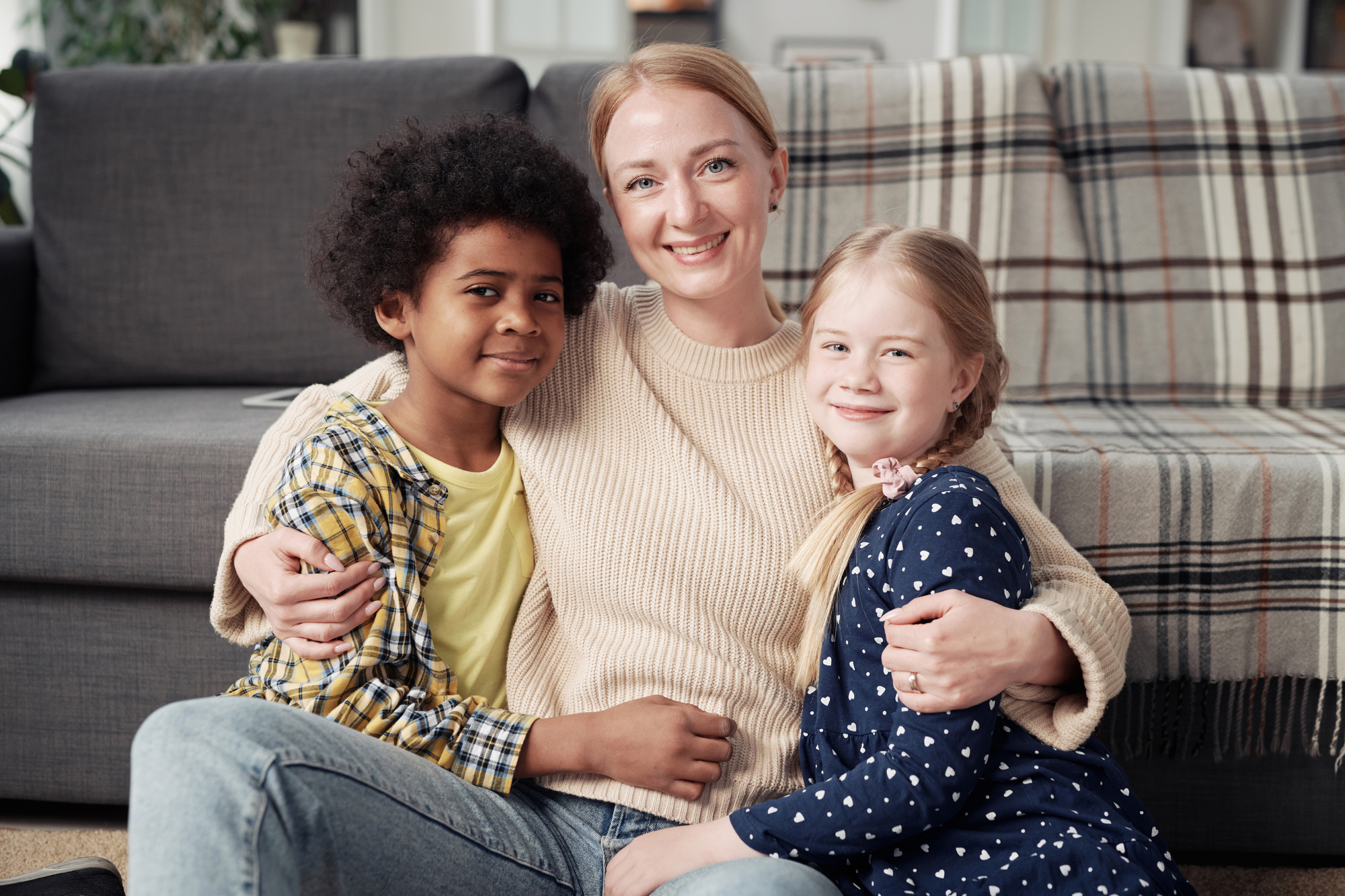 Woman sitting on the floor smiling and hugging two children, one with curly dark hair and the other with blonde hair, in a cozy living room setting.